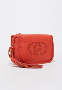 Červená kožená crossbody taška s textúrovaným povrchom, zlatými kovovými doplnkami, detailom s třásněmi a perforovaným dizajnom loga na prednej strane.