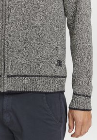 Cardigan lavorato a maglia grigio e nero con un motivo testurizzato, dotato di una toppa nera con logo sulla parte anteriore inferiore e rifiniture nere sui polsini e sull'orlo.
