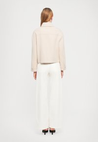 Giacca beige cropped con colletto e maniche lunghe. Indossata sopra pantaloni bianchi a gamba larga, che mostrano una texture liscia e un design semplice.
