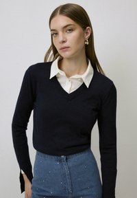 Marineblauwe v-hals trui van zachte stof, met lange mouwen en een aangesloten ontwerp, gelaagd over een witte blouse met kraag, gecombineerd met hoge taille denim.