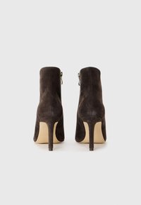Botines de ante marrón con un alto tacón de aguja, que cuentan con una cremallera lateral y una textura suave. Diseño simple y elegante, sin patrones visibles.
