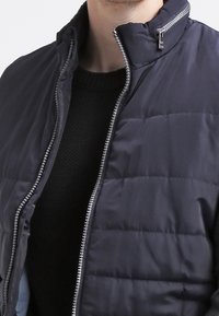 Granatowa pikowana kamizelka z kołnierzem stojącym i srebrnym zamkiem błyskawicznym. Posiada poziomą pikowaną teksturę i warstwowy design na czarnym dzianinowym swetrze.