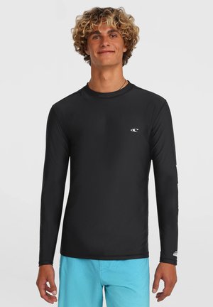 Jeune homme aux cheveux bouclés portant un rash guard noir à manches longues et un short de bain bleu clair, debout devant un fond uni.