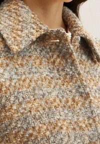 Close-up van een persoon die een textuurgegeven, meerkleurige wollen jas draagt met een grote kraag en knoopsgatdetail bij de nek.