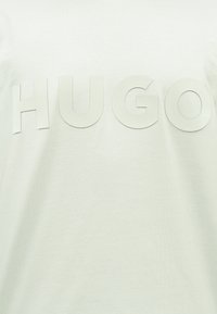 Λευκό ύφασμα με μεγάλα, ανάγλυφα γράμματα που σχηματίζουν τη λέξη «HUGO» σε μινιμαλιστική, μοντέρνα γραμματοσειρά στο κέντρο.