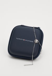 Navy blue Tommy Hilfiger zippered case with logo and a thin silver chain necklace featuring a circular emblem and a bar pendant.