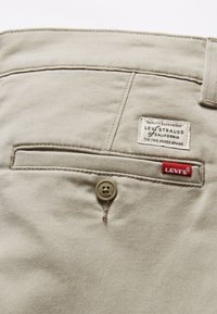 Pantaloni beige con una tasca posteriore, dotati di un bottone marrone e di un'etichetta rossa Levi's ben visibile. Il materiale ha una texture liscia, con dettagli di cucitura sottili.