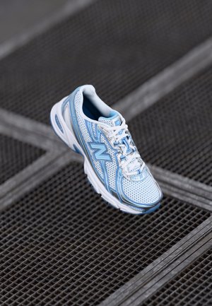 White and light blue athletic sneaker with mesh design floating above a dark grey metal grid surface.