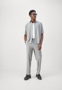 Lichtgrijs shirt met korte mouwen en knopen, gecombineerd met een bijpassende broek met trekkoord, beide met een geribde textuur. Zwarte schoenen en een zonnebril.