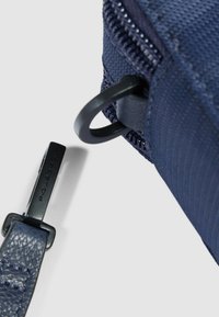 Esquina de una bolsa de tela azul marino con una cremallera brillante y una correa de cuero con una hebilla metálica grabada con "PIQUADRO".