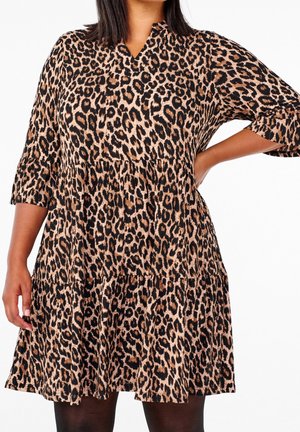 Femme portant une robe imprimé léopard longueur genou avec manches 3/4, debout avec une main sur la hanche sur fond blanc.