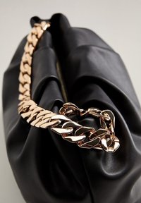 Sac en cuir noir avec une anse en chaîne dorée, présentant un design décontracté et une fermeture éclair visible sur le dessus. La chaîne a un maillon en forme de cœur.