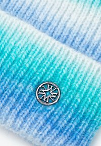 Gorro de punto con un gradiente ombre de verde a azul, con un detalle de logotipo circular plateado. La textura es suave y ligeramente peluda.