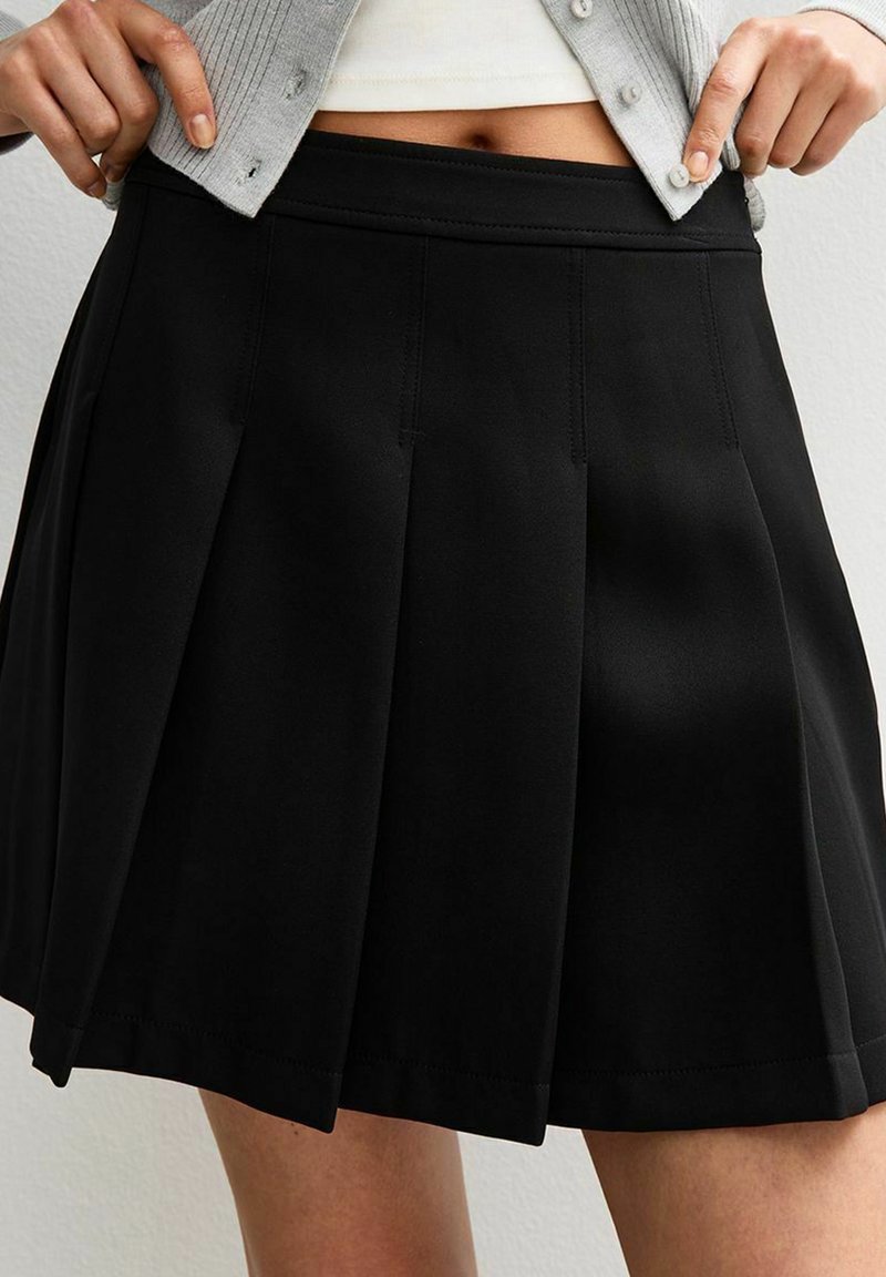 Zwarte geplooide rok met een gladde textuur, voorzien van een aansluitende tailleband en verticale stikdetails, eindigend boven de knie.