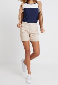 Shorts en coton beige avec un ourlet roulé, assortis à un haut à manches courtes bleu marine et blanc et à des baskets gris clair. Un léger cardigan est drapé sur les épaules.