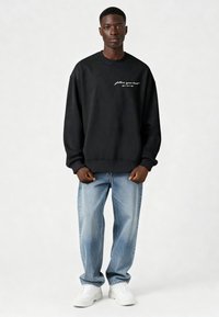 Jonge man staand, draagt een zwarte trui met tekst en pijl, lichtblauwe jeans en witte sneakers tegen een effen achtergrond.