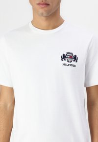 Hvit bomulls T-skjorte med rund hals og kort erm, med en brodert logo i marineblått og rødt på brystet.