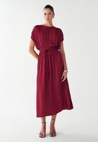 Vestido maxi burdeos con mangas cortas, cintura fruncida y cordón. Presenta una textura suave y llega a la mitad de la pantorrilla. Tacones negros con tiras.