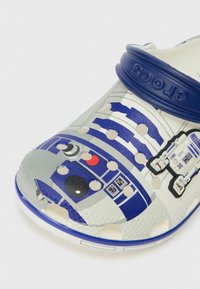 Otroški črno-beli klini Crocs s podobo R2-D2 iz Star Wars in modrim prilagodljivim trakom za peto z oznako Crocs.