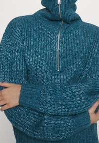 Pull en tricot teal avec un col montant et une fermeture éclair partielle, doté d'un motif texturé et de poignets côtelés. Matière en fil épais et doux.