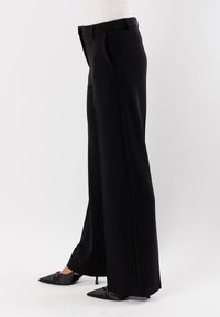 Pantaloni neri a gamba larga con una texture liscia, dotati di una vita alta e pieghe sottili, abbinati a scarpe nere a punta.