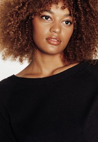 Maglione nero lavorato a maglia con scollo ampio. Il tessuto appare morbido e con una texture. I capelli sono ricci e voluminosi. Le labbra hanno una finitura lucida.