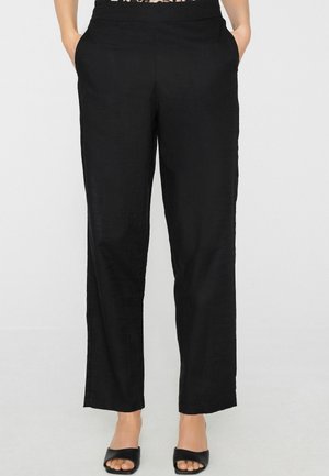 Pantalon droit noir avec poches latérales porté par une personne debout, associé à des talons ouverts noirs sur fond blanc.