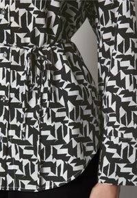Camicia a righe bianco e nero con un tessuto morbido e texture, dotata di una cintura in vita arricciata, bottoni e maniche lunghe con polsini.