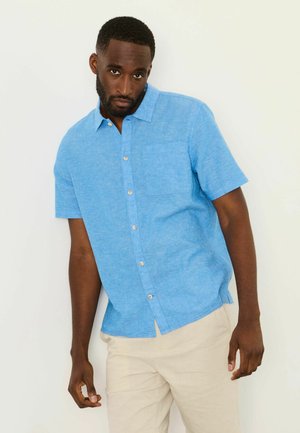 Mann trägt ein hellblaues Kurzarm-Hemd mit Knöpfen und beigen Hosen, steht vor einem schlichten hellen Hintergrund.