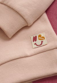 Weiches pinkfarbenes Strickgewebe mit geripptem Bündchen und einem kleinen aufgenähten Patch, das ein lächelndes Gesicht, ein Herz und die Buchstaben "NRPS" zeigt.