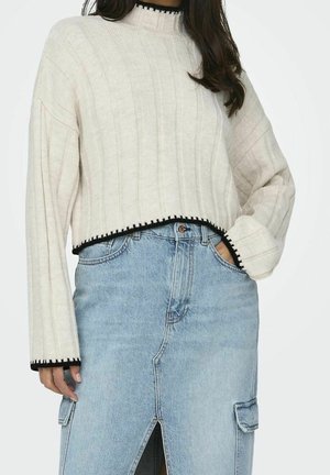 Mujer con suéter crema corto con ribete negro y falda vaquera azul claro con abertura frontal y bolsillos cargo, mano en el bolsillo.