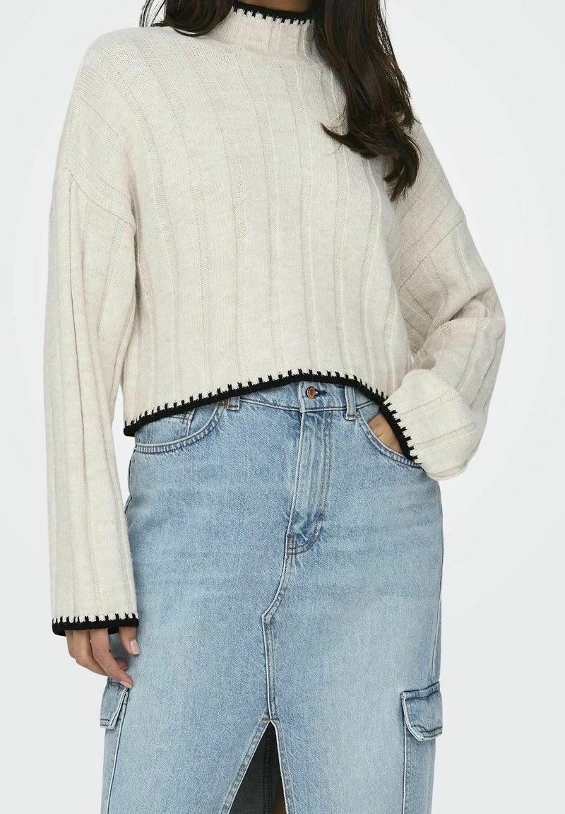 Femme portant un pull court crème avec une bordure noire et une jupe en jean bleu clair avec une fente devant et des poches cargo, main dans la poche.