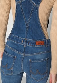 Dunkelblaue Jeanslatzhosen mit einem braunen, gestreiften Langarmoberteil. Merkmale sind ein Lederetikett, Taschen mit geschwungenen Nähten und ein taillierter Schnitt.