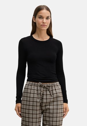 Donna con capelli castani lisci che indossa una maglia nera aderente a maniche lunghe e pantaloni marroni a quadri con coulisse, in piedi davanti a uno sfondo semplice.