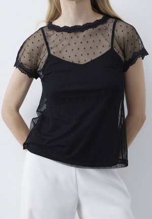 Kobieta ubrana w czarny przezroczysty top w groszki z krótkim rękawem, nałożony na czarny top na ramiączkach, w połączeniu z białymi spodniami na jednolitym tle.