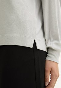 Hellegraues Langarmoberteil mit lockerer Passform, das an Saumseite einen Schlitz und einen glatten, strukturierten Stoff aufweist. Kombiniert mit schwarzen Hosen.