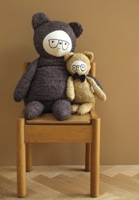 Grote bruine pluche beer met een bril en een kleinere beige beer met een strikje, beide zittend op een houten stoel tegen een beige muur.