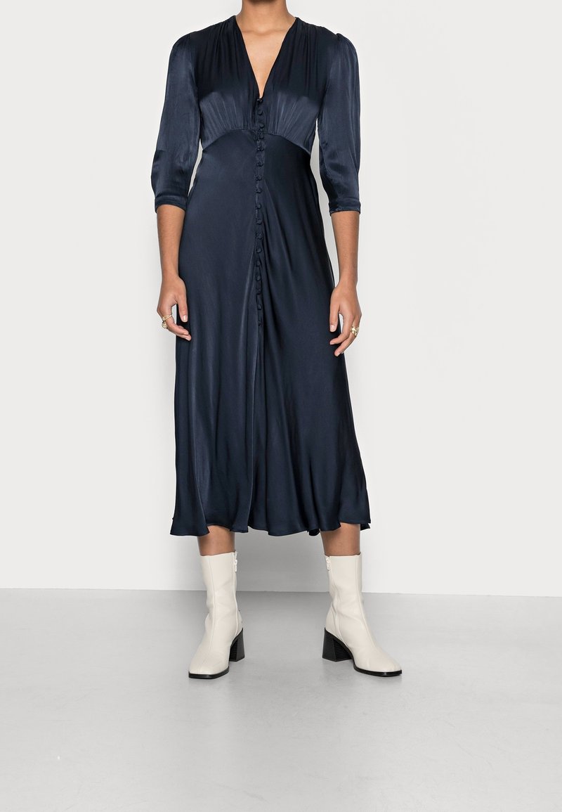 Robe midi en satin marine avec un décolleté en V profond, six boutons sur le devant, manches trois-quarts et une silhouette fluide, associée à des bottines blanches.