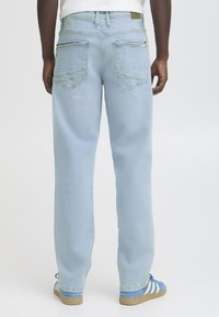 Moški nosi svetlo modre ravne jeans hlače in modre športne copate z belimi črtami, stoji pred enostavno ozadje.