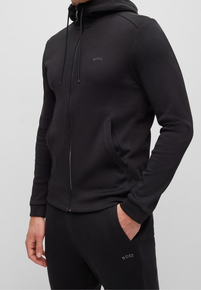 Man som bär en svart zip-up hoodie och matchande sweatpants med "BOSS"-logotyp, står med en hand i fickan mot en grå bakgrund.