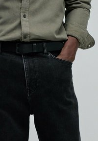 Persona vestida con una camisa de botones verde oliva y jeans negros, con una mano en el bolsillo; se puede ver un cinturón negro en los jeans.