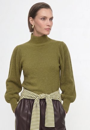Kvinde med kort brunt hår iført guld øreringe i ringform, en olivengrøn rullekravesweater og mørkebrune læderbukser med et stribet bælte.