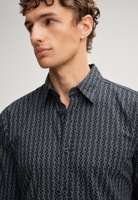 Camicetta nera con bottoni, caratterizzata da un motivo geometrico in tonalità più chiara, con colletto classico e tessuto testurizzato.