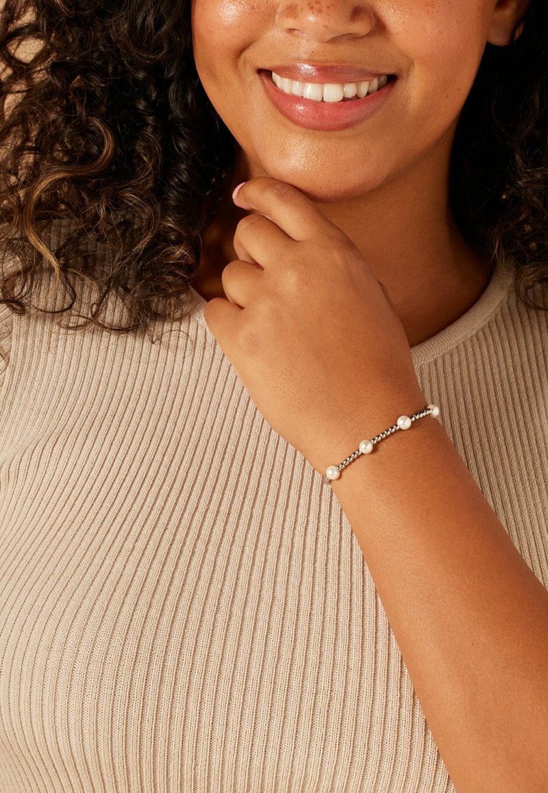 Zilveren armband met ronde witte parels, afgewisseld met kleine zilveren kralen. De hand van het model rust op de kin en toont het ontwerp van de armband.