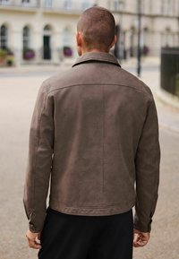 Brown suede jacket with a smooth texture, featuring a classic collar and buttoned cuffs, viewed from the back against a street background.