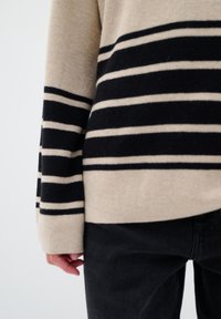 Beige en zwarte gestreepte trui met een losse pasvorm, gebreide manchetten en een zachte textuur. Gecombineerd met donkere zwarte jeans, gedeeltelijk zichtbaar.