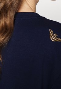 Marinblå tygtröja med en texturerad gul strassaccent i form av en vinge på den övre ryggen. Slät textur, enkelt mönster.