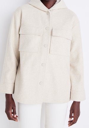 Chaqueta beige con capucha y cierre de botones a presión, dos grandes bolsillos delanteros con solapa, llevada sobre pantalones blancos, mostrando el torso y los brazos de una persona.