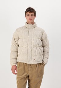 Pufferjakke i lys beige, tekstureret stof med høj krave. Har quiltede sektioner og et lille logo-patch. Parret med fløjl bukser.