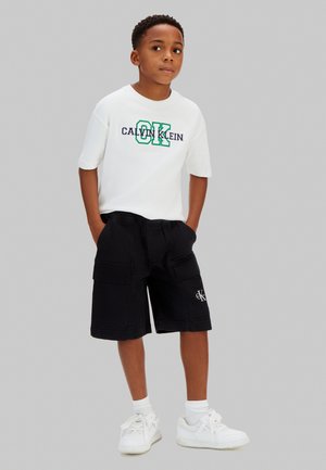 Junger Junge steht mit den Händen in den Taschen, trägt ein weißes Calvin Klein T-Shirt, schwarze Cargo-Shorts, weiße Socken und weiße Turnschuhe vor grauem Hintergrund.